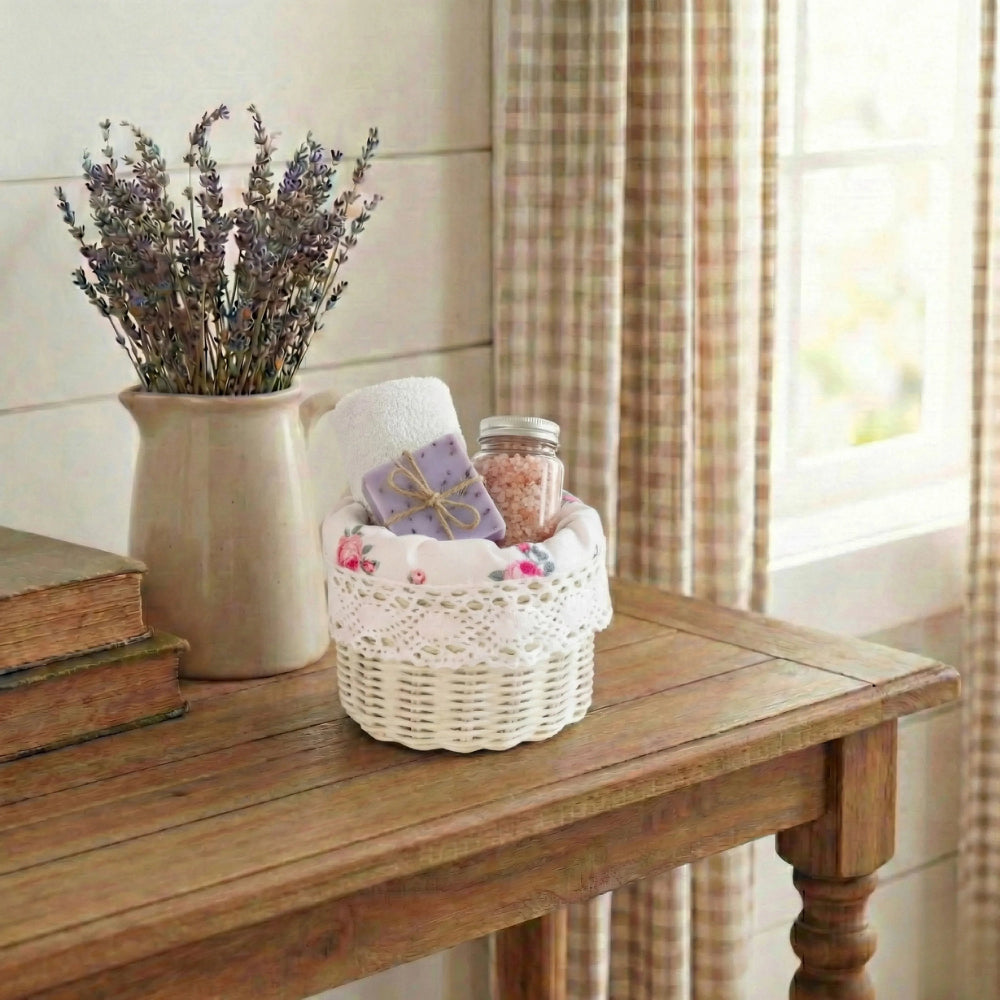 White mini wicker basket holding soap, bath salts, and a towel on a wooden table next to a lavender pitcher.