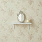 Ornate white oval mirror and a candle on a white shelf against floral rose wallpaper.