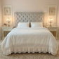 White ruffled bedding set on a bed with a grey tufted headboard, two white nightstands, and lit candelabra lamps.