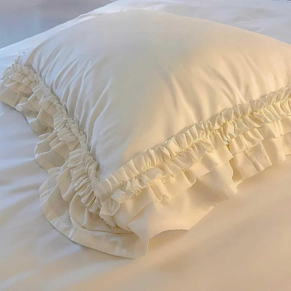 Close-up of a white ruffled pillowcase edge showing layered pleats resting on a smooth white bed sheet.