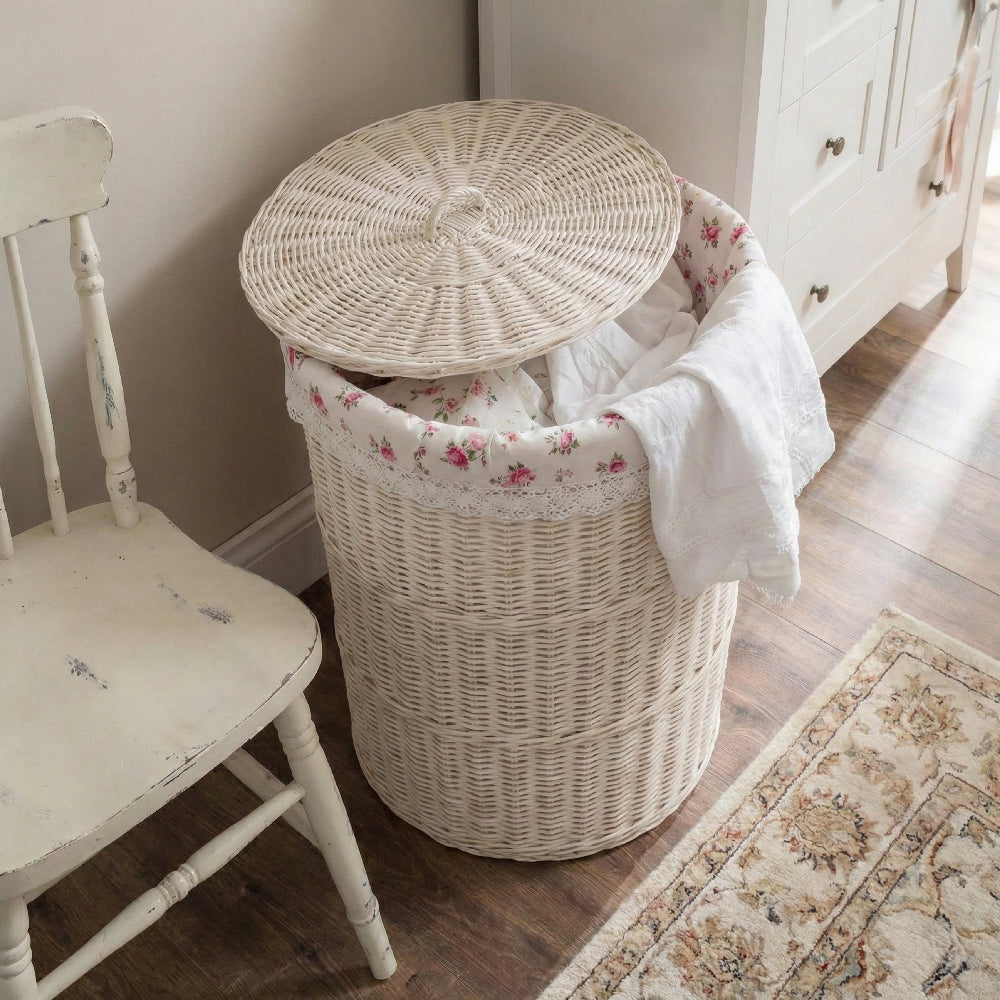 White wicker laundry basket with floral lining and lid next to a distressed white chair and a patterned rug.