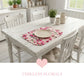 White wooden dining table with floral placemat and chairs in a bright room.