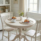 Breakfast for two with croissants and coffee on woven placemats at a white pedestal table with matching wooden chairs.