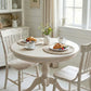 Round wooden dining table with chairs set for breakfast in a bright room.
