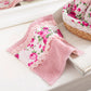 A pink face towel with a floral fleur print and lace detailing, displayed on a white surface next to a wicker basket with a pink and white floral design.