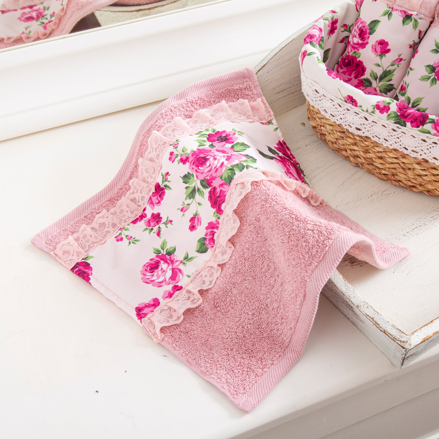 A pink face towel with a floral fleur print and lace detailing, displayed on a white surface next to a wicker basket with a pink and white floral design.