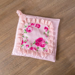 Square pink pot holder with a rose floral center, lace ruffled trim, and a hanging loop on a wooden surface.